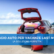 Auto rossa parcheggiata su una spiaggia con il bagagliaio aperto e pieno di valigie, pronta per una vacanza. In primo piano una valigia rossa. Testo sovrapposto: 'Noleggio auto per vacanze last minute: la soluzione perfetta'